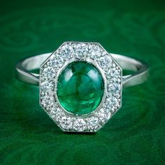 A glamorous Art Deco emerald diamond cluster ring from the 1920s crowned with a natural cabochon emerald in the centre, weighing approx. 1.6ct. It has a vivid green hue and inner glow that's complemented by sixteen bright brilliant cut diamonds around the border with SI1 clarity - H colour, approx. 0.80ct total. Emerald is the birthstone of May. The Romans considered it to be the sacred stone of the goddess Venus and was said to preserve love and provide hope. Its natural green colouring also ma Platinum Cluster Emerald Ring, Green Platinum Emerald Ring With Cluster Shape, Art Deco Diamond Rings With Cabochon, Art Deco Diamond Cabochon Rings, Cluster Green Emerald Ring In Platinum, Green Emerald Cluster Ring In Platinum, Classic Cabochon Emerald Ring, Art Deco Oval Emerald Ring With Brilliant Cut, Classic Diamond Cabochon Ring