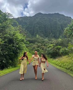 three women walking down the road in front of some trees and mountains, holding hands