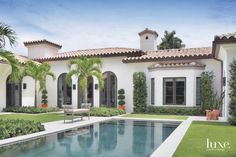 a large pool in front of a white house with palm trees and shrubs around it