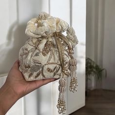 Add a touch of elegance and sophistication to your bridal look with our exquisite Cream Velvet Potli Bag. Crafted from premium velvet, this potli bag is a testament to timeless beauty and unmatched craftsmanship. The soft cream shade exudes a subtle charm that complements any bridal attire, while the intricate hand-embroidered zardozi and sequins work create a mesmerizing display of artistry. Each stitch and sequin is meticulously placed, showcasing the skill and dedication of our artisans. Designed for comfort and style, this potli bag features a stunning handmade braided handle adorned with delicate beads, offering both durability and a unique aesthetic. The handle is carefully crafted to ensure a comfortable grip, making it an ideal accessory for the bride on her special day. To further Elegant Silver Embroidered Bags, Embellished Beige Evening Bag As Gift, Beige Pouch Potli Bag For Evening, Beige Embellished Evening Bag As Gift, Elegant Cream Bridal Accessories, Luxury Pearl Embroidered Potli Bag For Party, Elegant Potli Bag With Pearl Handle, Evening Potli Bag With Pearl Handle, Traditional Evening Clutch With Pearl Handle