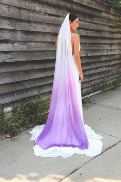 a woman in a purple and white wedding dress standing on the sidewalk next to a wooden wall