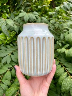 a hand holding a ceramic vase in front of green leaves