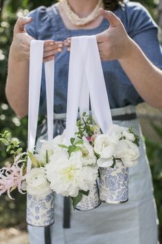 a woman holding three vases with flowers in them and ribbons hanging from the handles