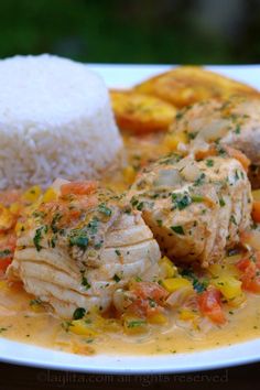 a white plate topped with chicken and vegetables covered in sauce next to rice on top of a wooden table