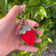 a hand holding a tiny crocheted strawberry keychain in front of green leaves