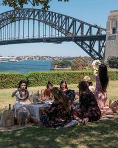 group of women sitting on the grass in front of a bridge eating and drinking wine