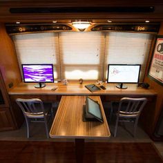 two computer monitors sitting on top of a wooden desk in front of a window with blinds