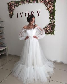 a woman standing in front of a floral wall wearing a white dress with long sleeves