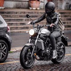 a man riding on the back of a motorcycle down a cobblestone street next to a parked car