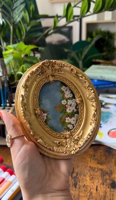 a person holding up a small gold frame with flowers on it in front of some plants