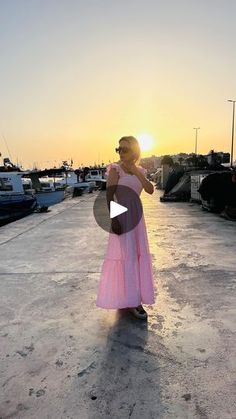 a woman in a pink dress is standing on the dock with boats at sunset behind her