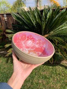 a hand holding a pink and green bowl in front of a palm tree on the grass