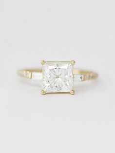 a gold ring with a princess cut diamond in the center, on a white background