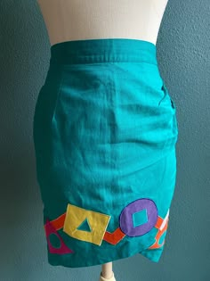 Vintage Skirt in Bright Teal Green with multi color geometric graphic shapes appliquéd on  -skirt is tulip shaped  -zips and buttons in the back  -waist has elastic in part of band  -Made by Deborah Lunt Boutique  Measurements laying flat: waist 12.5 inches / stretched 14 inches  Length 22.5" Retro Fitted Mini Skirt With Pockets, Retro Fitted Patchwork Skirt, Green Patchwork Mini Skirt, Fitted Retro Cotton Mini Skirt, Fitted Retro Green Mini Skirt, Green Fitted Retro Mini Skirt, Retro Fitted Skirt With Pockets, Spring Retro Fitted Pencil Skirt, Retro Fitted Pencil Skirt For Spring