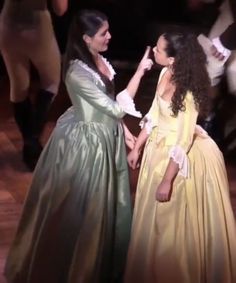 two women in long dresses standing next to each other on a wooden floor with dancers behind them