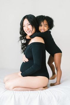 two women in black bodysuits sitting on a white bed and posing for the camera