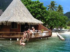 two people sitting on a dock in the water next to a hut with thatched roof