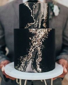 a black and gold wedding cake with white frosting on the top is being held by a man in a gray suit