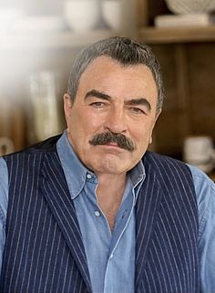 a man with a moustache wearing a blue shirt and striped vest is looking at the camera