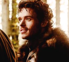 a man with curly hair wearing a fur coat and looking at another man in the woods