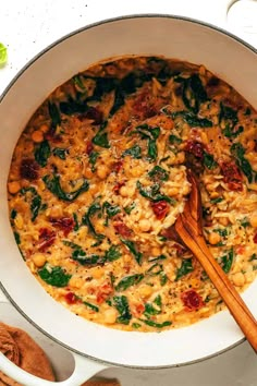 a white pot filled with pasta and spinach on top of a table next to a wooden spoon