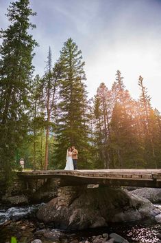 a bride and groom standing on a bridge over a river in the woods at sunset