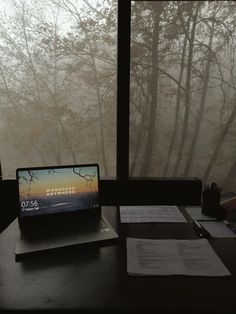 a laptop computer sitting on top of a desk next to a window with trees outside