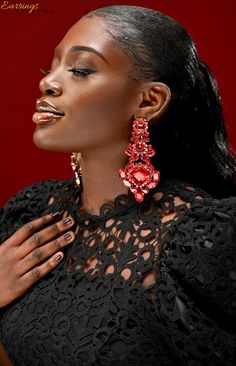 a woman with long black hair wearing red earrings and a black lace top, standing in front of a red background