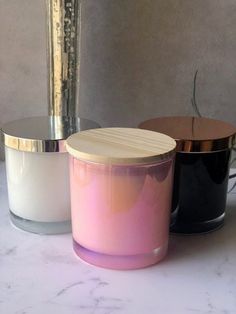 three different colored candles sitting next to each other on a white counter top with a metal vase in the background