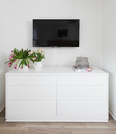 a flat screen tv mounted on the wall above a white dresser with flowers in vases