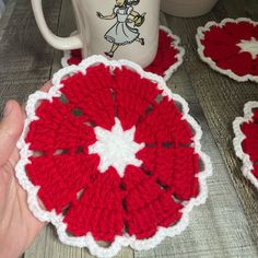 a hand holding a crocheted coaster next to two coffee mugs on a table