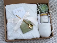 an open box containing candles, mugs and other items on a blanket with a white ribbon