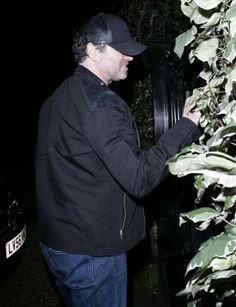 a man standing next to a black clock in front of some bushes and trees at night