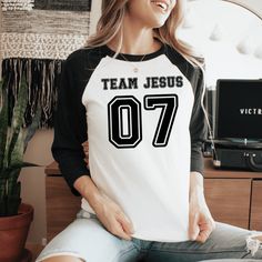 This Team Jesus Jersey Shirt is the perfect Christian shirt for any sports fan! It's a sporty stylish way to share your faith and heart for Jesus with anyone! Plus it's super soft and comfy. Whether your heading to your favorite sports game, running errands or a weekend get away, this cute Team Jesus shirt is a comfy casual Jesus shirt for any occasion. A stylish spin on the classic baseball raglan. The combed cotton blend makes it super soft, comfortable, and lightweight. You don’t even have to Black Tops With Lettering For Sports Events, Black Collegiate Tops With Lettering, Team Spirit Tops With Lettering For Game Day, Varsity Sports Top With Lettering, Black Sporty Tops With Lettering, Sporty Black Tops With Lettering, Sporty Tops With Lettering For Game Day, Sporty Tops With Lettering For Sports Season, Sporty Tops With Lettering For Sports