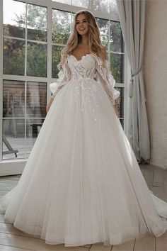 a woman standing in front of a window wearing a white wedding dress with long sleeves