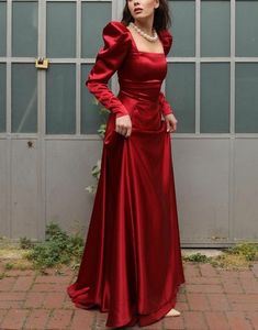 a woman in a long red dress is standing by a wall and posing for the camera