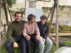 three people are sitting on a swing in the grass and one person is smiling at the camera