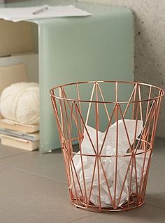 a copper wire trash can on the floor next to some books and yarn ball holders