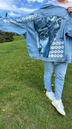 a woman is standing in the grass holding up a jean jacket with an image of a man's face on it