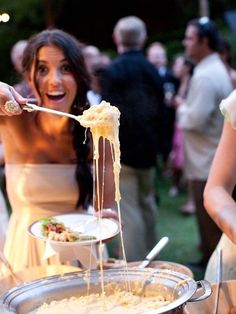 a woman is eating spaghetti from a silver dish on a table with other people in the background