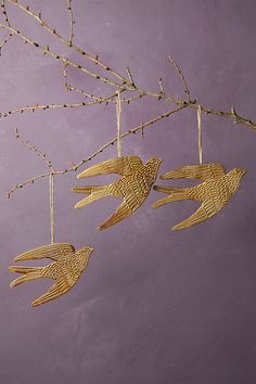 three gold bird ornaments hanging from a branch on a purple wall with branches in the background