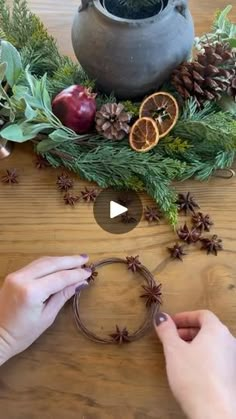 two hands are making a wreath with pine cones and oranges on the table next to a potted plant