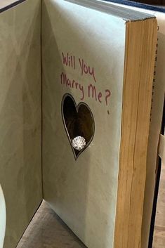 an open book with a diamond ring in the middle and will you marry me? written on it