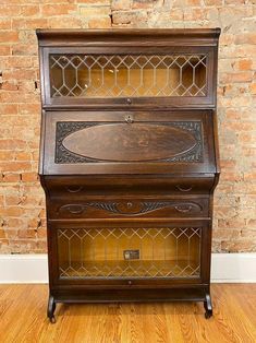 an old wooden desk with glass doors on the top and bottom shelf, in front of a brick wall