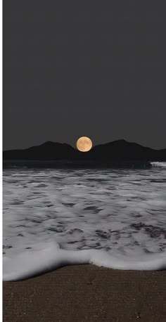 a full moon is seen over the ocean with waves coming in to shore and mountains in the distance