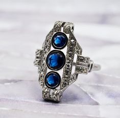 This exquisite vintage anti-tarnish rhodium plated ring is meticulously crafted from 925 solid sterling silver. It is adorned with three sapphire stones as its centerpiece, which are complemented by sparkling cubic zirconia accent stones. The dimensions of the ring are notable, with the sapphire arrangement measuring 0.90 inches in length and 0.65 inches in width, creating a striking and elegant presence on the finger. This piece is a testament to classic style and timeless craftsmanship. Stampe Sapphire Stones, Plated Ring, Sapphire Stone, Multi Stone Ring, Multi Stone, Birthday Present, Birthday Presents, Stone Rings, Rhodium Plated