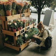 a woman sitting on the sidewalk next to a shelf filled with potted plants and flowers