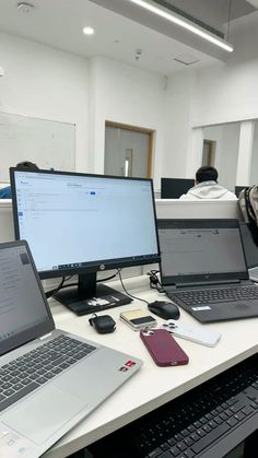 three laptops are sitting on a desk in an office with people working behind them