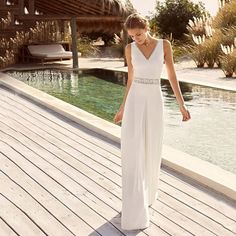 a woman in a white dress is standing on a wooden deck near the water and looking down