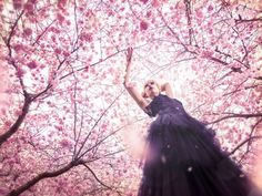 a woman in a black dress is standing under pink flowers and trees with her hands up